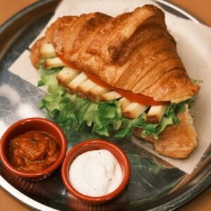 Croissant sandwich with cheese, lettuce, and tomato, served with red bowls of sauce and creamy dip.